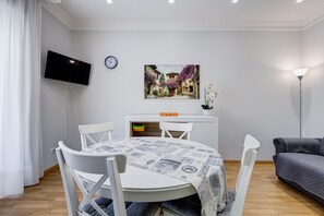 A dining area with a round table, white chairs, a flat-screen TV mounted on the wall, a clock, and a framed picture above the table. - Vatican Flat—2 Bedrooms, 2 En-suite Bathrooms, and a Balcony (Rome)