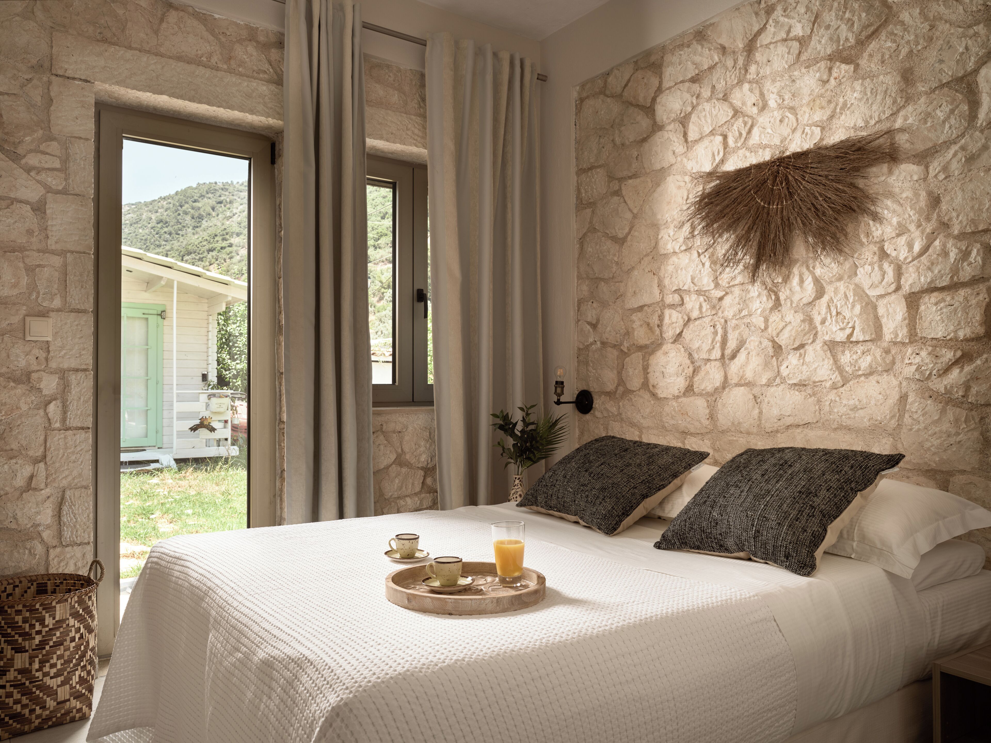 Een slaapkamer met een stenen muur, een bed met witte lakens, twee kussens, een nachtkastje met een glas sap, en een dienblad met kopjes.