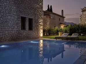 Ein Steinhaus mit Pool, Liegestühlen und einem gut beleuchteten Garten im Abenddämmerung.