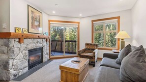 Ein Wohnzimmer mit Kamin aus Stein, einer Couch, einem Sessel und Blick ins Freie durch große Fenster.
