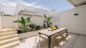 En terrasse med et bord og stole, omgivet af planter og et hvidt gelænder.