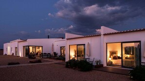 A row of modern, white buildings with large windows and doors, a gravel driveway, and outdoor seating areas.