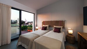 A hotel room with two beds, a balcony, a view of a park, and a lamp on a side table.