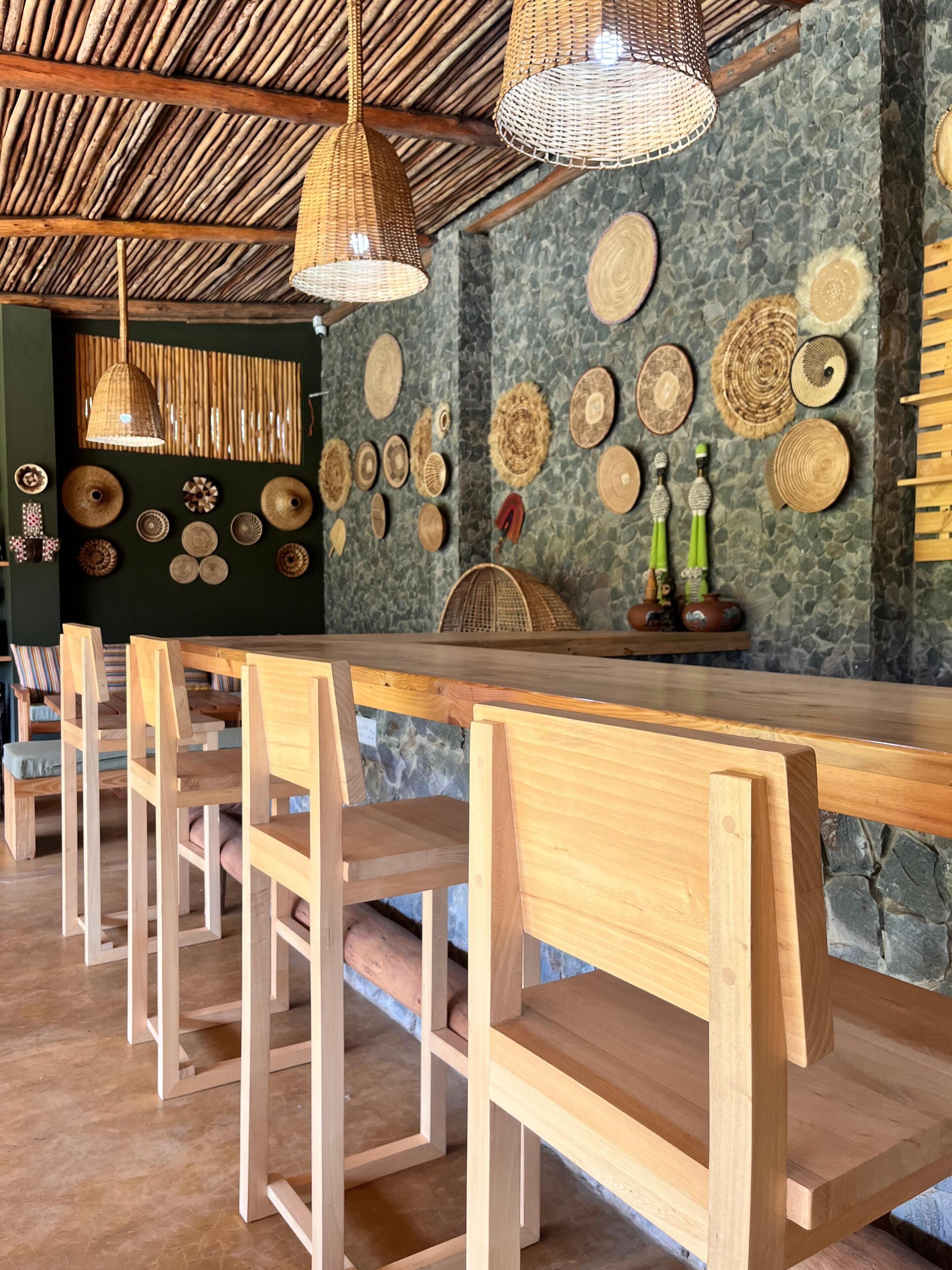 A wooden bar area with woven light fixtures, a stone wall with woven mats, and a row of wooden chairs.