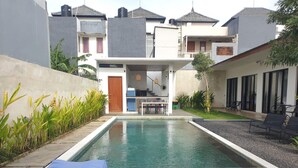 Una piscina con terraza de madera, zona de comedor y pequeño jardín con plantas.