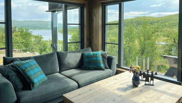 A room with a gray sofa, a wooden coffee table, and a view of a lake and forest through large windows. - 360-degree views from a mountain top in Numedal (Rødberg)