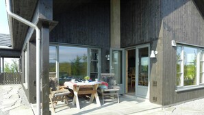A modern building with large glass doors, a stone patio, and a wooden table with chairs. - 360-degree views from a mountain top in Numedal (Rødberg)