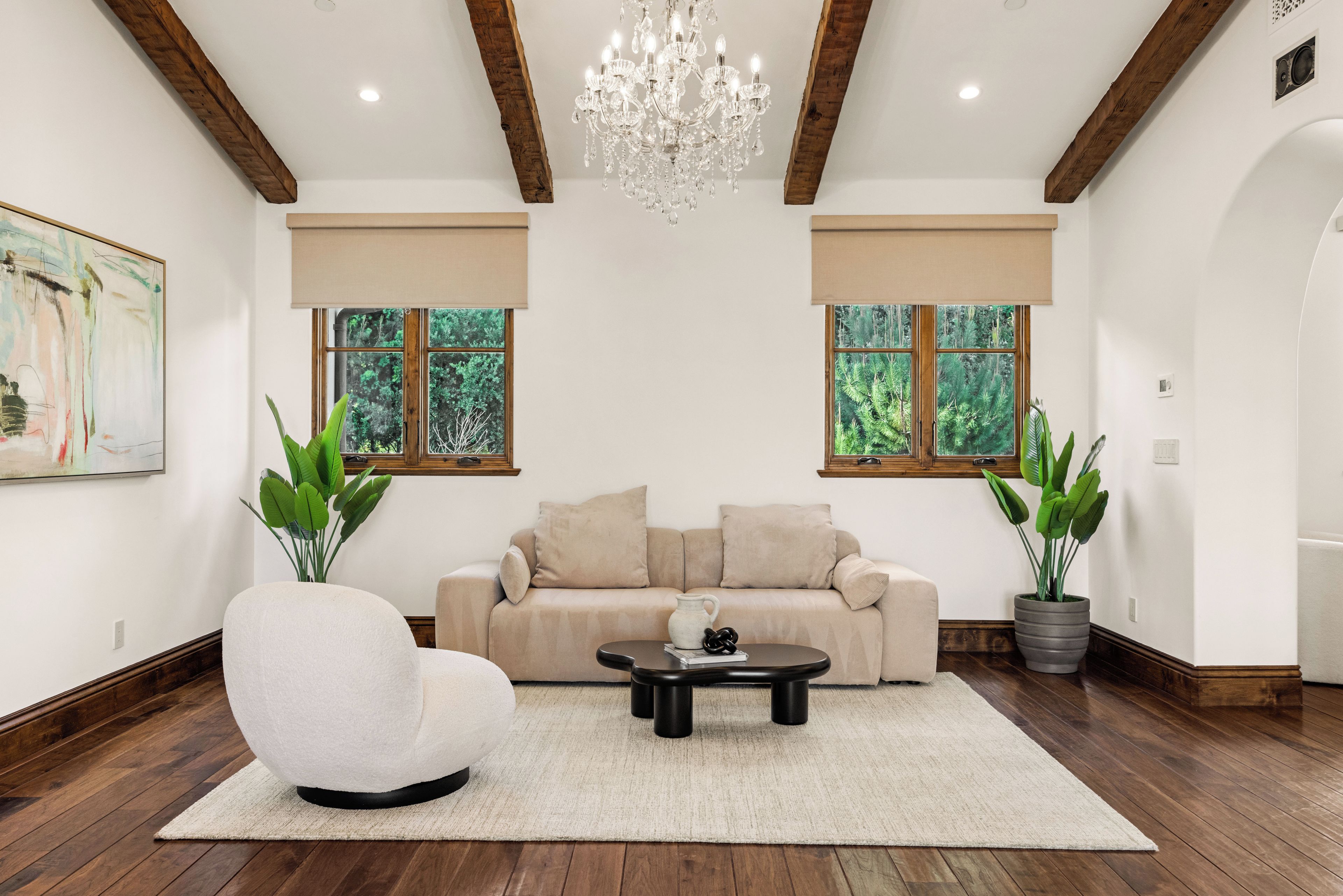 A spacious living room with a beige sofa, a white armchair, a round coffee table, and two large windows with a view of greenery.