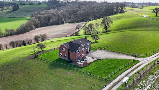 A rural house with a fenced yard, surrounded by green fields and trees.
