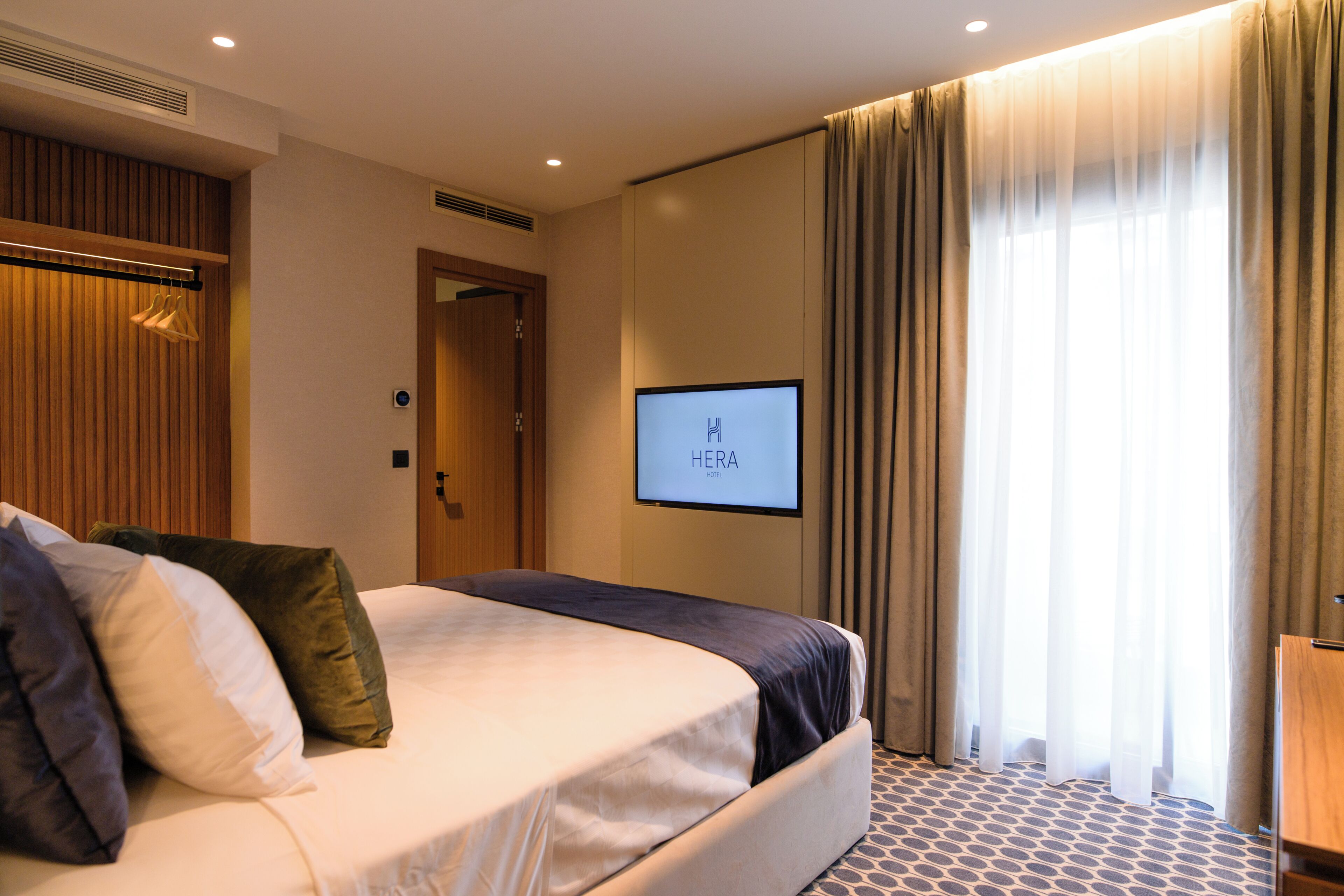 A hotel room with a bed, a television mounted on the wall, and a window with curtains.
