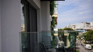 A balcony with two chairs, a glass railing, and a view of a city street. - Hera Hotel Axd (Alexandroupoli)