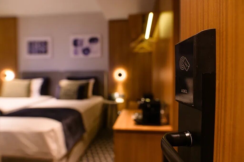 Habitación de hotel con cama, escritorio con cafetera y puerta con panel de control.