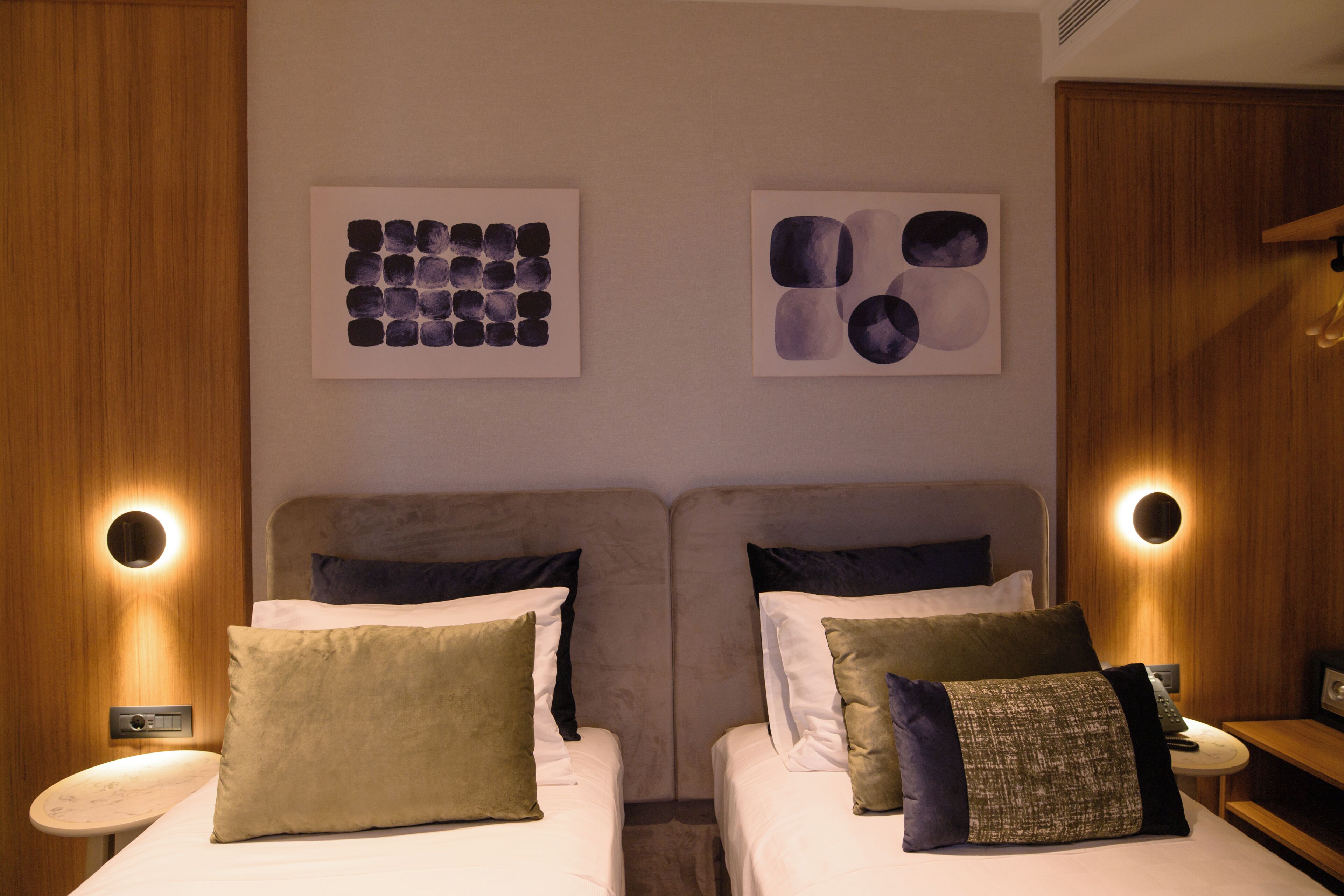 A hotel room with two beds, each with a pillow and a bedside table. There are wall-mounted light fixtures on either side of the beds.