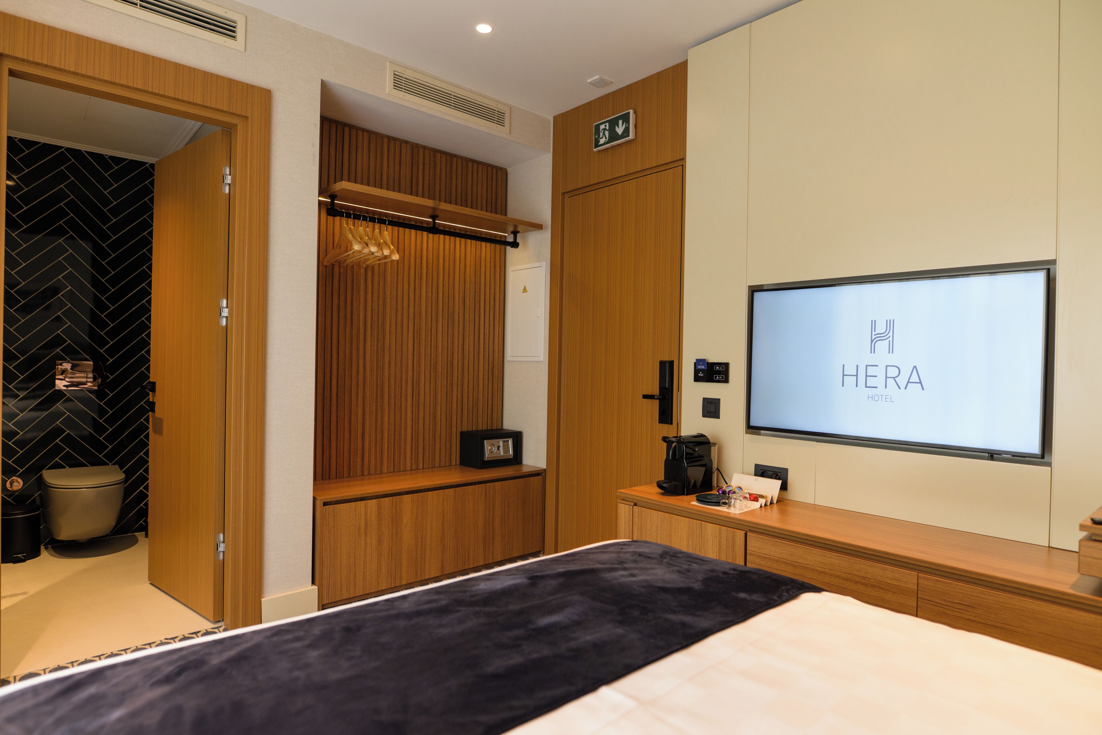 A hotel room with a bed, a TV displaying 'HERA HOTEL', a wardrobe, a coat rack, and a small table with a coffee maker.