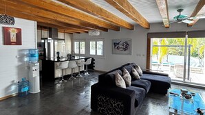 Amplia sala de estar con piso oscuro, un sofá modular negro, una mesa de centro de vidrio y una cocina al fondo.