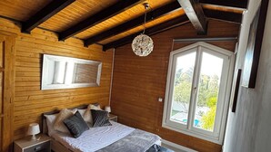 Un dormitorio con techo de madera, una cama con reposabajeras, una mesita de noche, una lámpara de pared y una ventana con vistas.