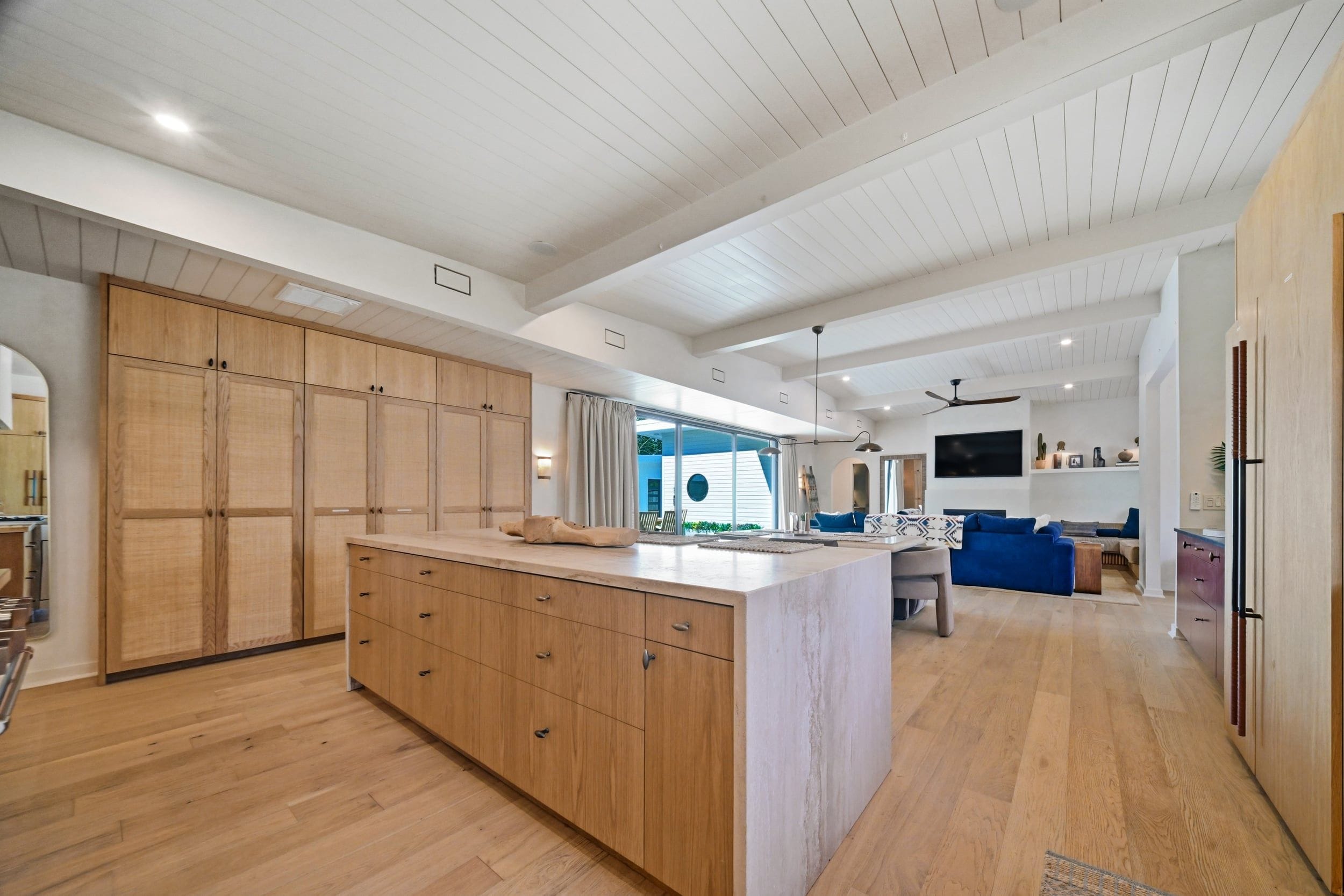 A spacious kitchen with wooden cabinets and a central island, leading to a living area with a blue sofa and a flat-screen TV.