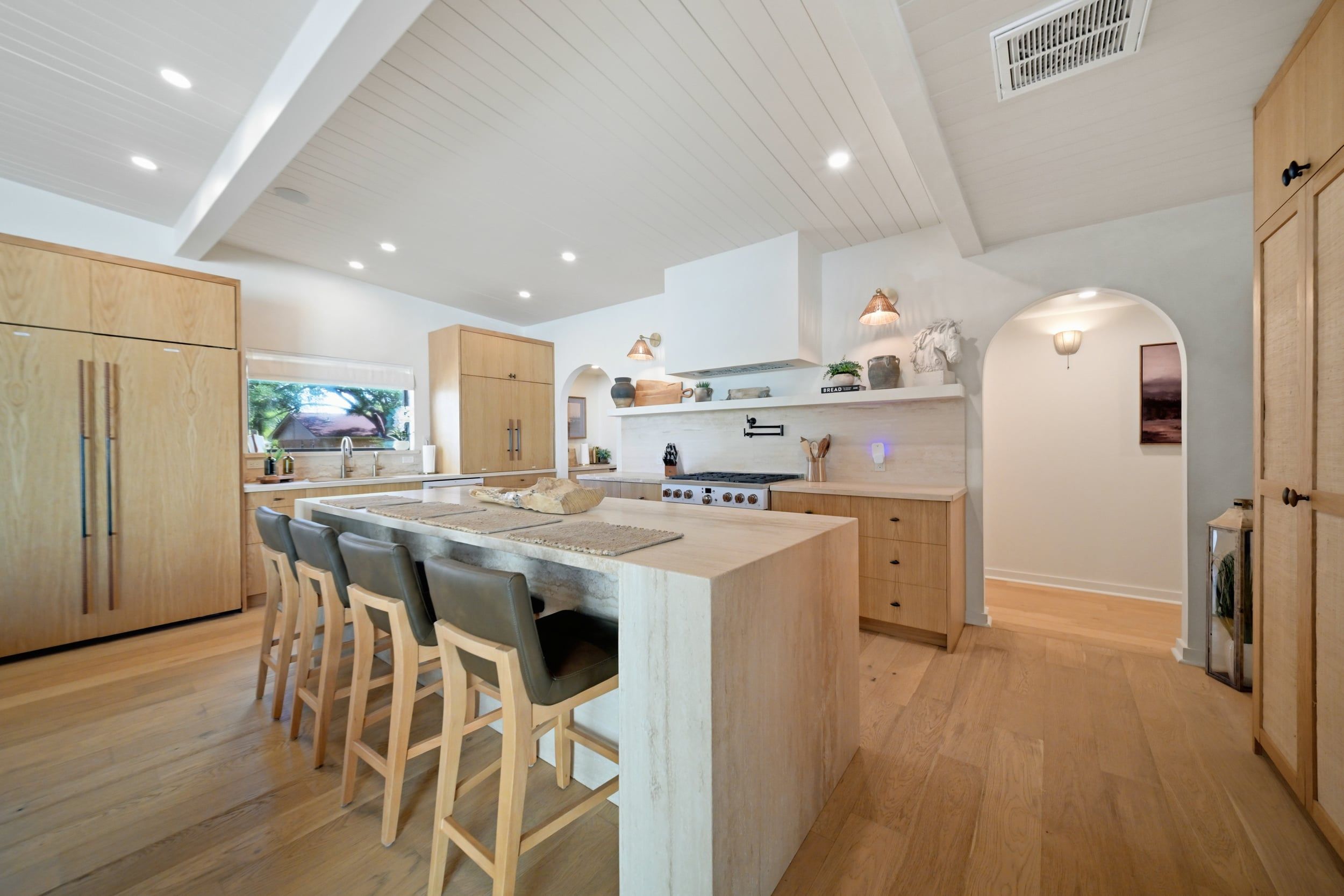 A modern kitchen with a central island, bar stools, wooden cabinets, and a view of the outdoors.