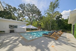 A swimming pool with lounge chairs and umbrellas, surrounded by trees and a modern house. - Vine Retreat Near Texas A&M (Bryan)