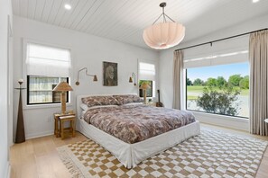 Ein Schlafzimmer mit einem großen Bett, Blick auf eine grüne Landschaft, einer Pendelleuchte und einem Gemälde an der Wand.
