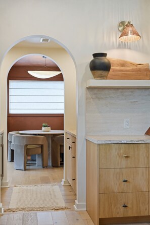 A modern kitchen with an arched doorway leading to a dining area, wooden cabinets, a hanging light fixture, and a potted plant on the counter. - Vine Retreat Near Texas A&M (Bryan)