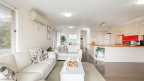 Un salon moderne avec un canapé blanc, une table basse et un coin cuisine doté de meubles blancs et d’un mur d’accent rouge.