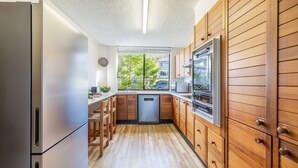 A compact kitchen with wooden cabinets, a stainless steel refrigerator, and a window with a view of greenery. - The Dunes 5, 38 Marine Dr - pool, tennis court, Wi-Fi (Fingal Bay)