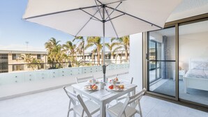 A balcony with a table set for breakfast, a large umbrella, and a view of palm trees and buildings. - The Dunes 5, 38 Marine Dr - pool, tennis court, Wi-Fi (Fingal Bay)