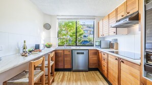 A compact kitchen with wooden cabinets, a dishwasher, and a window with a view of greenery and buildings. - The Dunes 5, 38 Marine Dr - pool, tennis court, Wi-Fi (Fingal Bay)