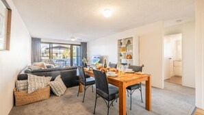 A living room with a sofa, dining table, chairs, and a balcony with a view. - The Dunes 5, 38 Marine Dr - pool, tennis court, Wi-Fi (Fingal Bay)