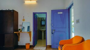 A room with a purple door labeled 'ROOM NO. 115', a wooden cabinet, a small table with a mirror, and two orange chairs.