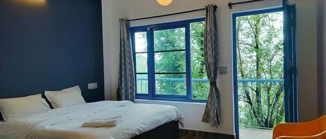 A bedroom with a bed, a window with curtains, a hanging light fixture, and a view of greenery outside.