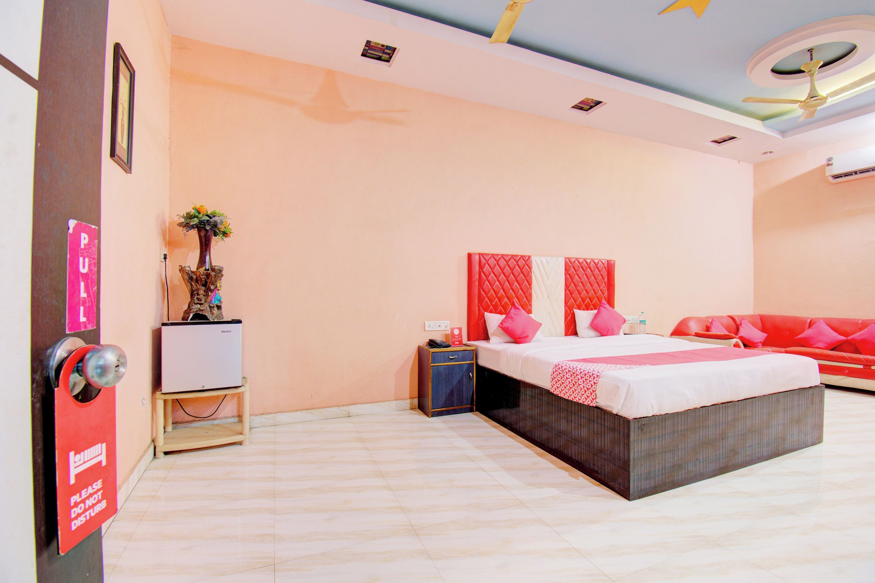 Una habitación de hotel con cama, un sofá rojo, una mesita con un jarrón y un televisor montado en la pared.