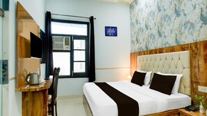 A hotel room with a bed, a desk with a kettle, a chair, a window with curtains, and a wall with a blue sign. - Deluxe Chahal Residency (Amritsar)