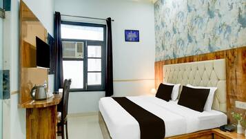 A hotel room with a bed, a desk with a kettle, a chair, a window with curtains, and a wall with a blue sign.