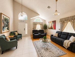 A spacious living room with a high ceiling, a large window, and a view of the outdoors. There are two sofas, a coffee table, and a painting on the wall.