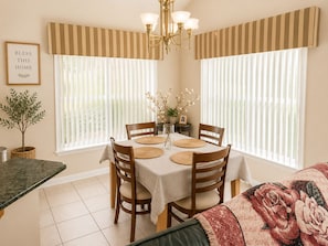 A dining area with a table set for four, surrounded by chairs, a chandelier, and large windows with blinds.