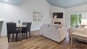 A modern living room with a dining area, a sofa, a TV mounted on the wall, and a large window.