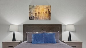 A bed with a quilted cover, two bedside lamps, a headboard, and a cityscape painting on the wall.