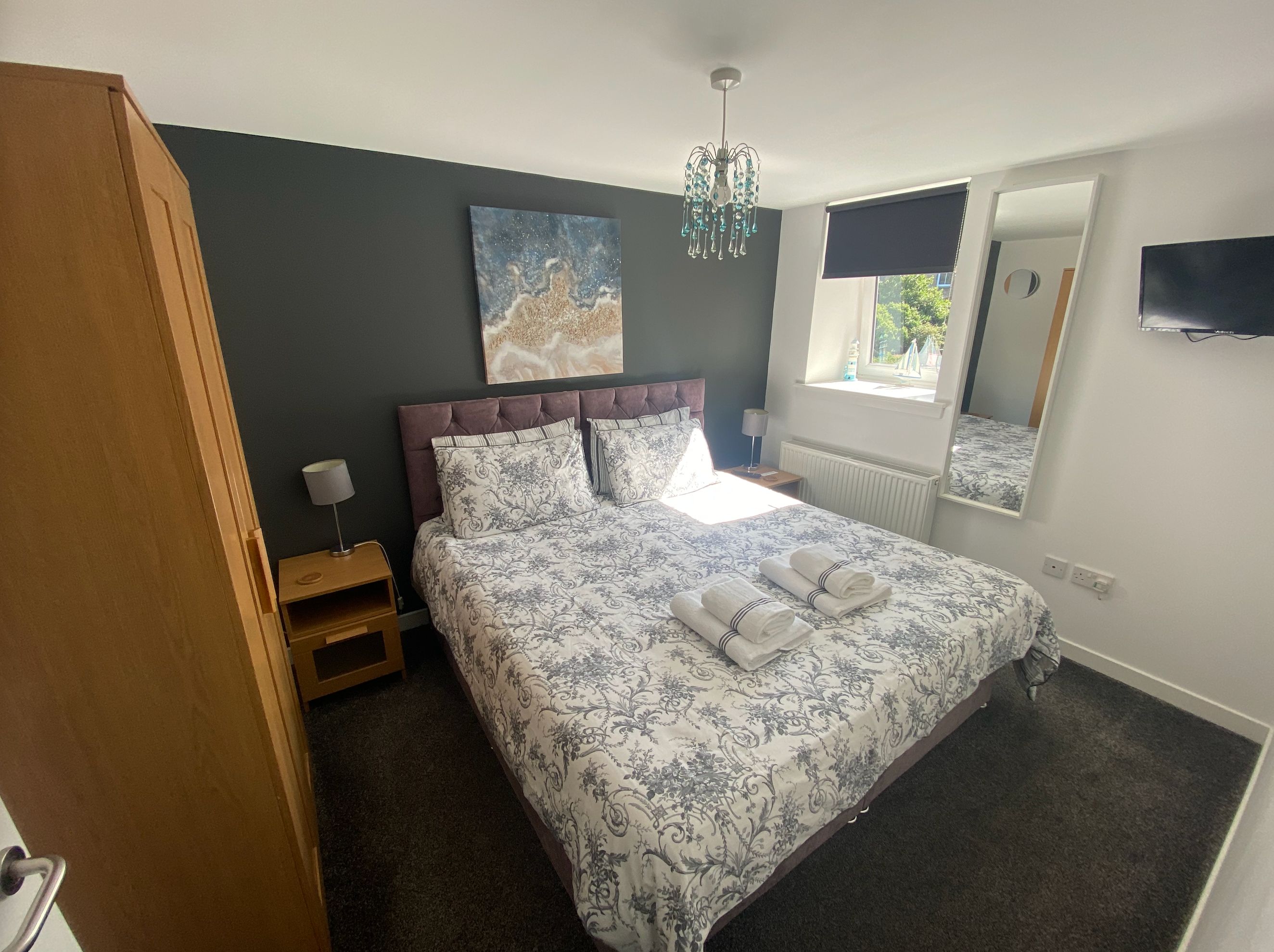 Une chambre à coucher avec un lit, une table de chevet, une lampe, une fenêtre, un radiateur et un tableau accroché au mur.