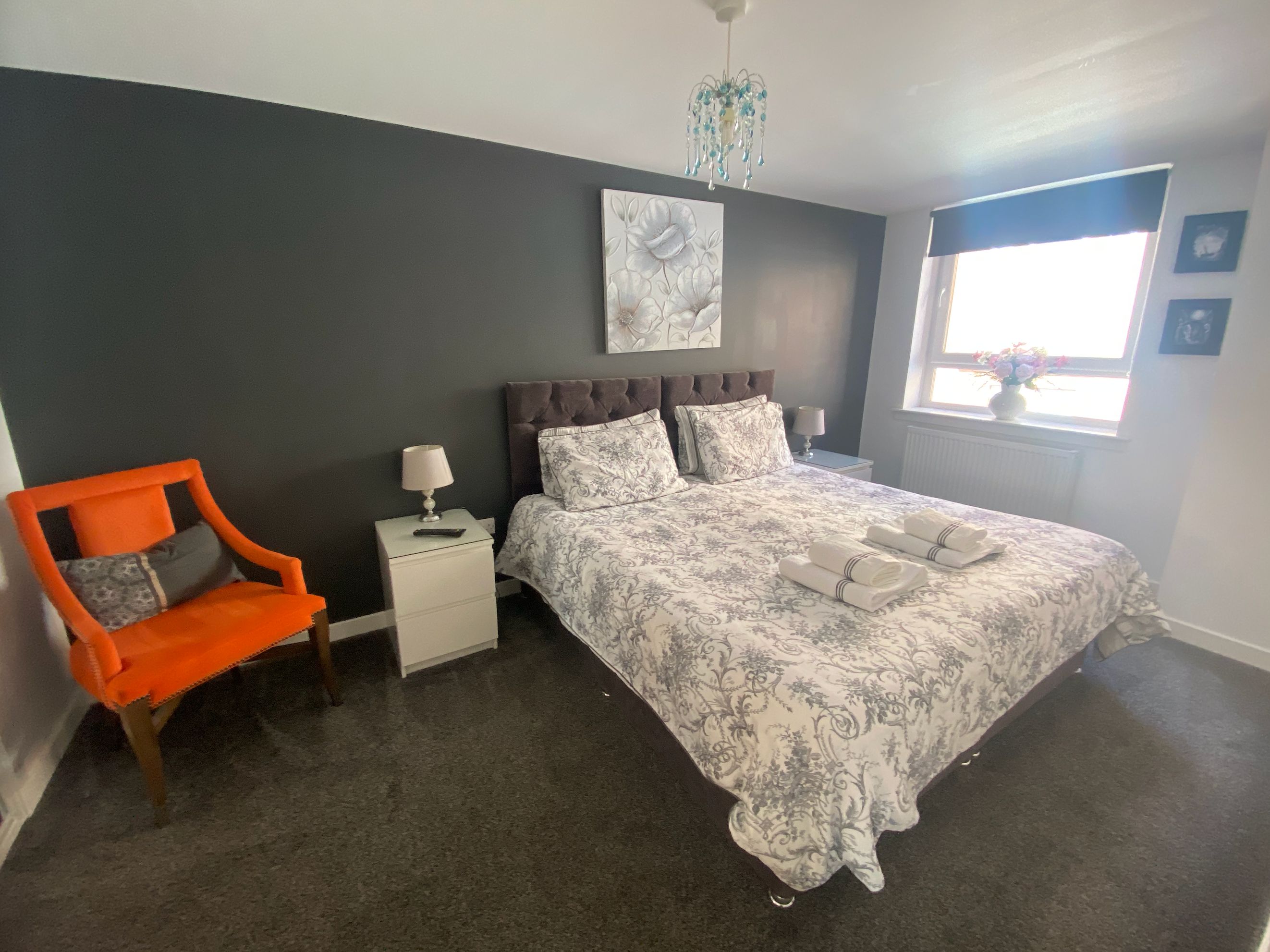 Quarto com cama, mesa de cabeceira, cadeira, abajur, lustre e janela com vista.