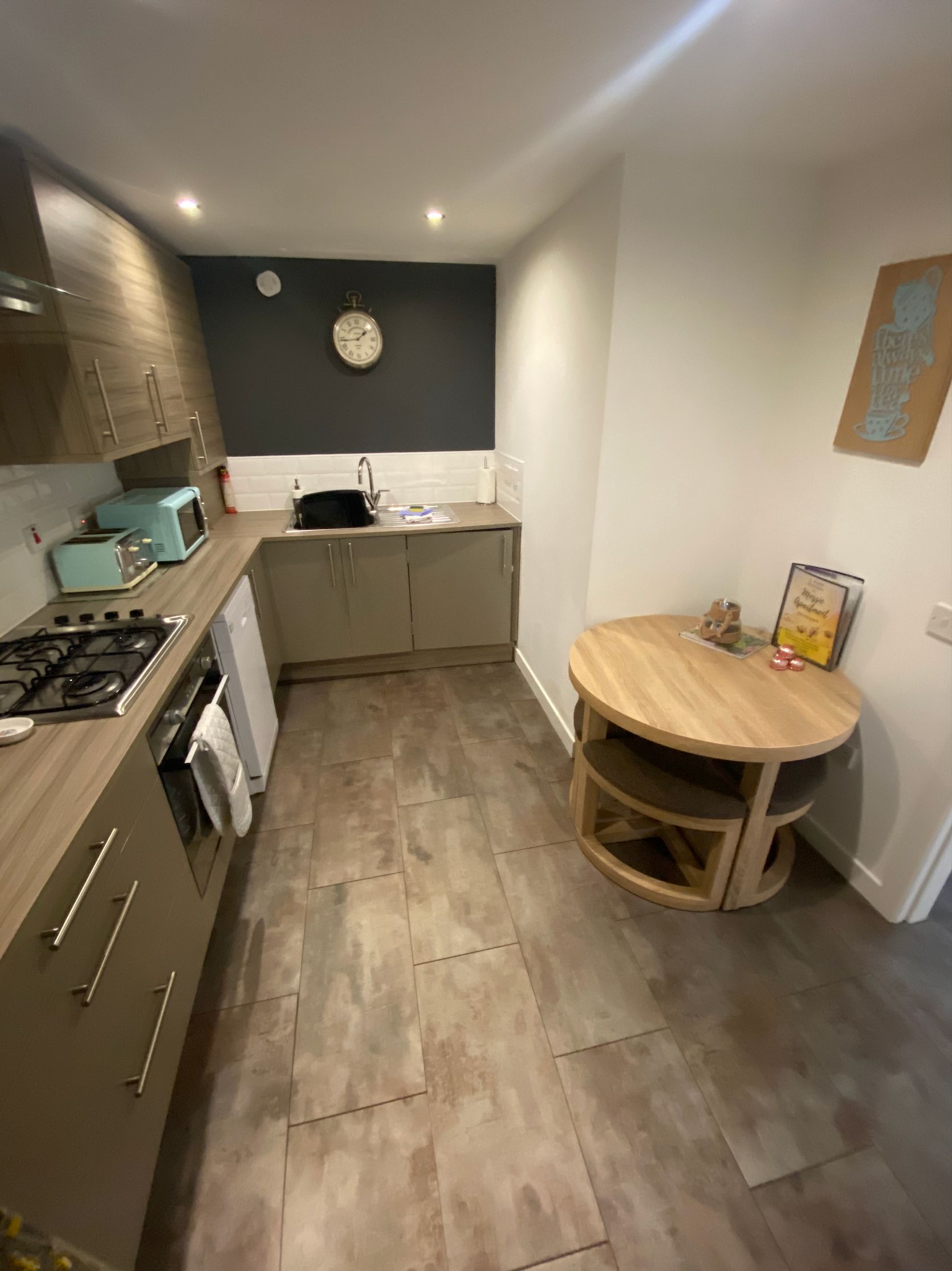 A compact kitchen with a gas stove, a sink, and a microwave. There's a round wooden table with chairs and a clock on the wall.