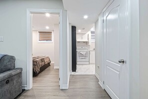 A narrow hallway leading to a bedroom with a bed and a gray sofa, a kitchen area with white cabinets and appliances, and a white door on the right. - Bright Basement Suite w Parking Near Mclennan Park (Kitchener)