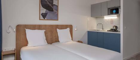A double bed with white linens, a wooden headboard, a small bedside table, a wall-mounted lamp, a kitchen area with a microwave, and a blue cabinet.