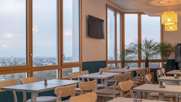 Een restaurant met meerdere tafels en stoelen, grote ramen met uitzicht op de stad en een aan de muur bevestigde televisie.