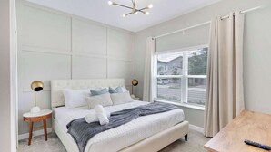 A bedroom with a large bed, a bedside table, a lamp, a window with curtains, and a view of the outside. - ChampionsGate 5BR | Pool, Xbox + Oasis Resort (Davenport)