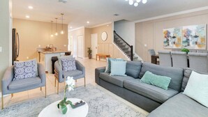 A modern living room with a grey sofa, armchairs, a coffee table, and a staircase leading to an upper level. - ChampionsGate 5BR | Pool, Xbox + Oasis Resort (Davenport)