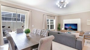 A modern living room with a dining table, sofa, TV, and large windows. - ChampionsGate 5BR | Pool, Xbox + Oasis Resort (Davenport)