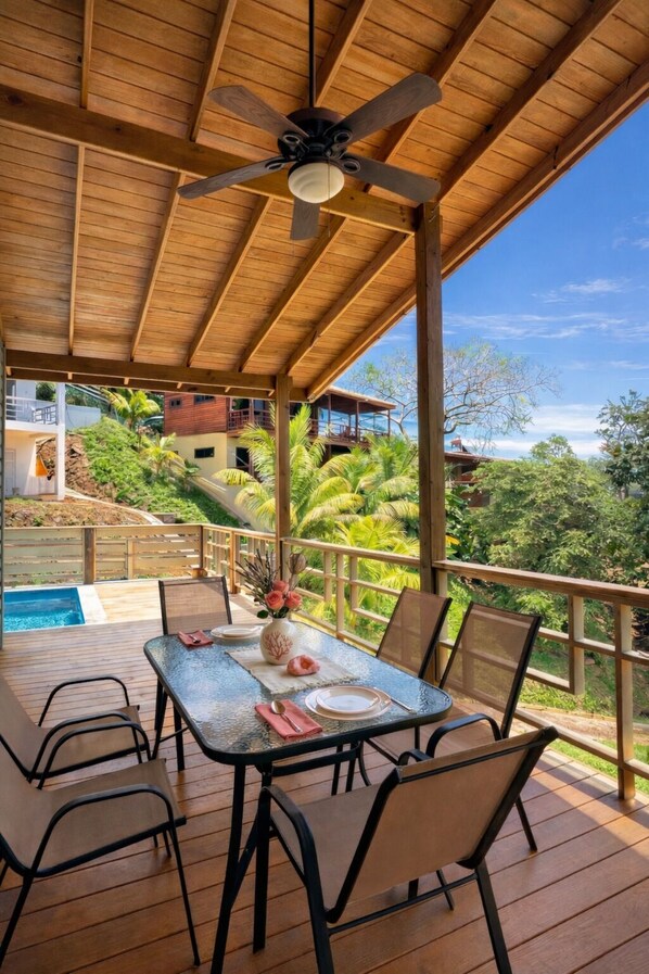 Eine Holzterrasse mit Tisch und Stühlen, einem Deckenventilator und Blick auf ein Schwimmbad und Gebäude im Hintergrund.