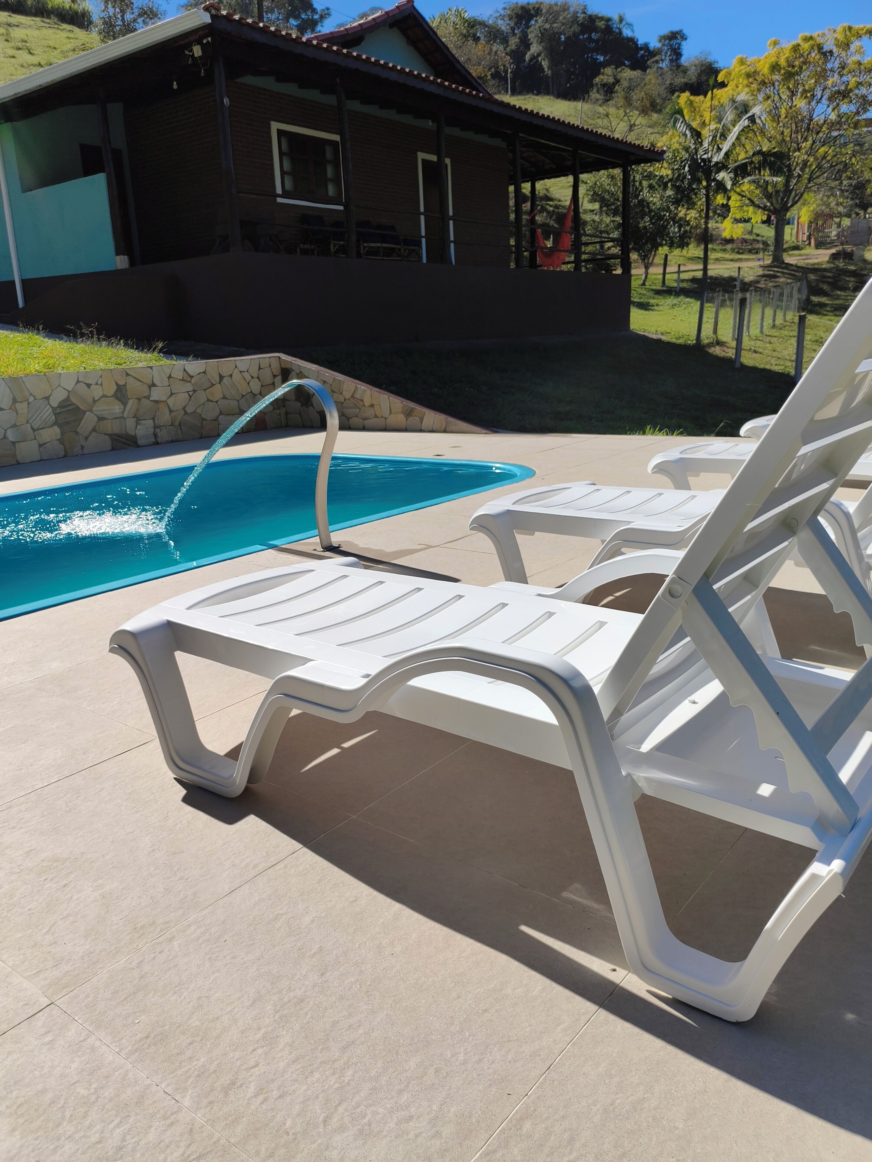 A pool area with white lounge chairs, a stone wall, and a house in the background.
